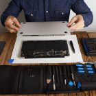 service man opens backside topcase cover computer laptop before repairing cleaning fixing it with his professional tools from toolkit box near wooden table front view scaled